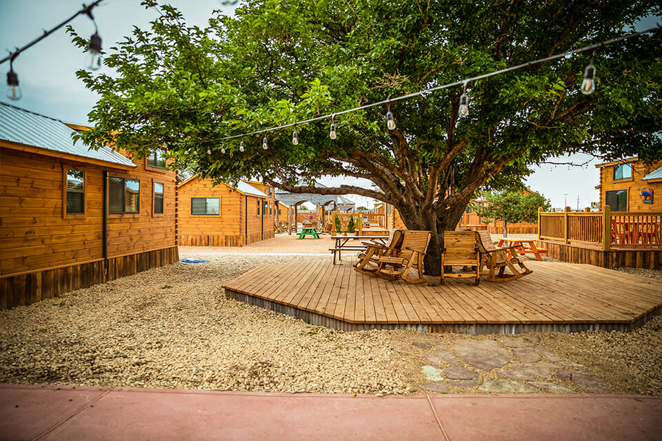 Outdoor wooden patio area with chairs, tables, and a large tree in a rustic setting.