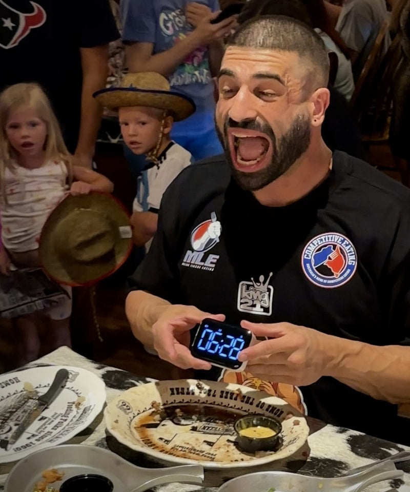 Man in a black shirt with his mouth wide open while holding a digital clock after completing the 72oz steak challenge