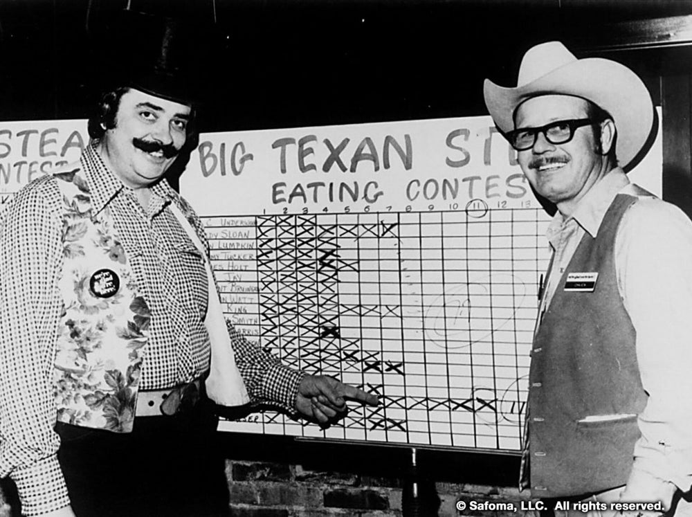 Two men standing in front of a 'Big Texan Steak Ranch' eating contest sign.
