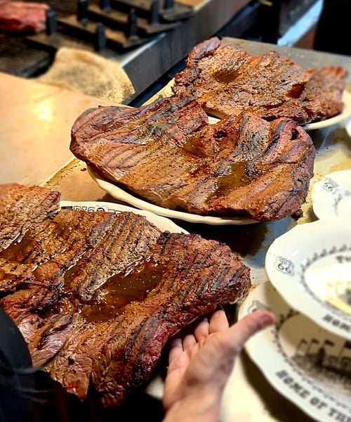 three large grilled beef steaks on a plate with a hand holding one of the plates, in a kitchen setting.