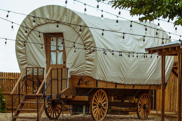 Wooden covered wagon with a white canvas cover, surrounded by string lights and wooden structures.