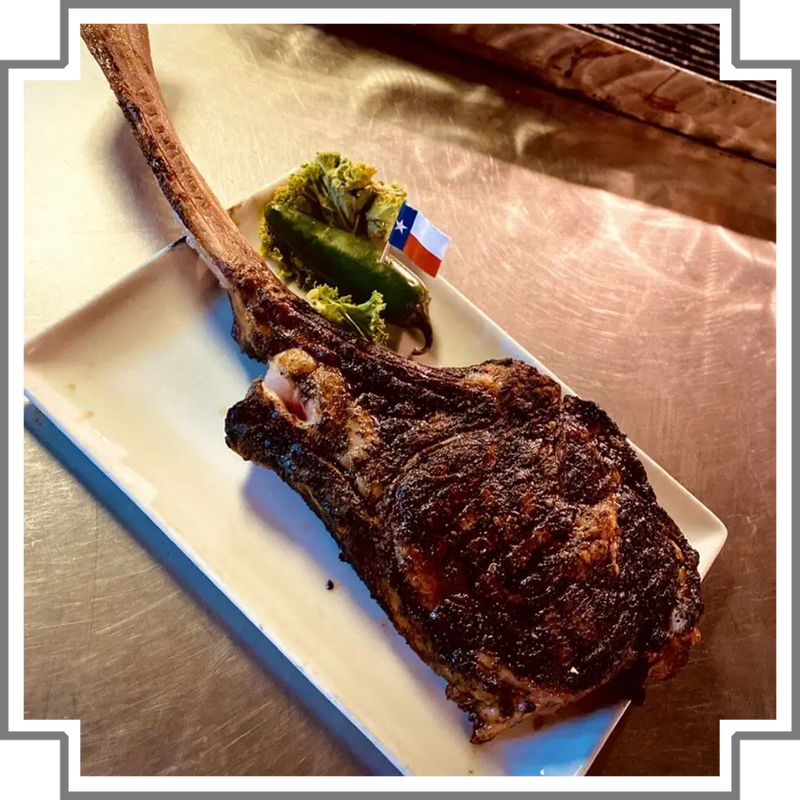 Grilled tomahawk steak on a white plate with a small flag and garnish, on a metallic surface.