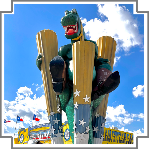 Large statue of a dinosaur with cowboy boots and Texas flags against a blue sky with the Big Texan building in the background.