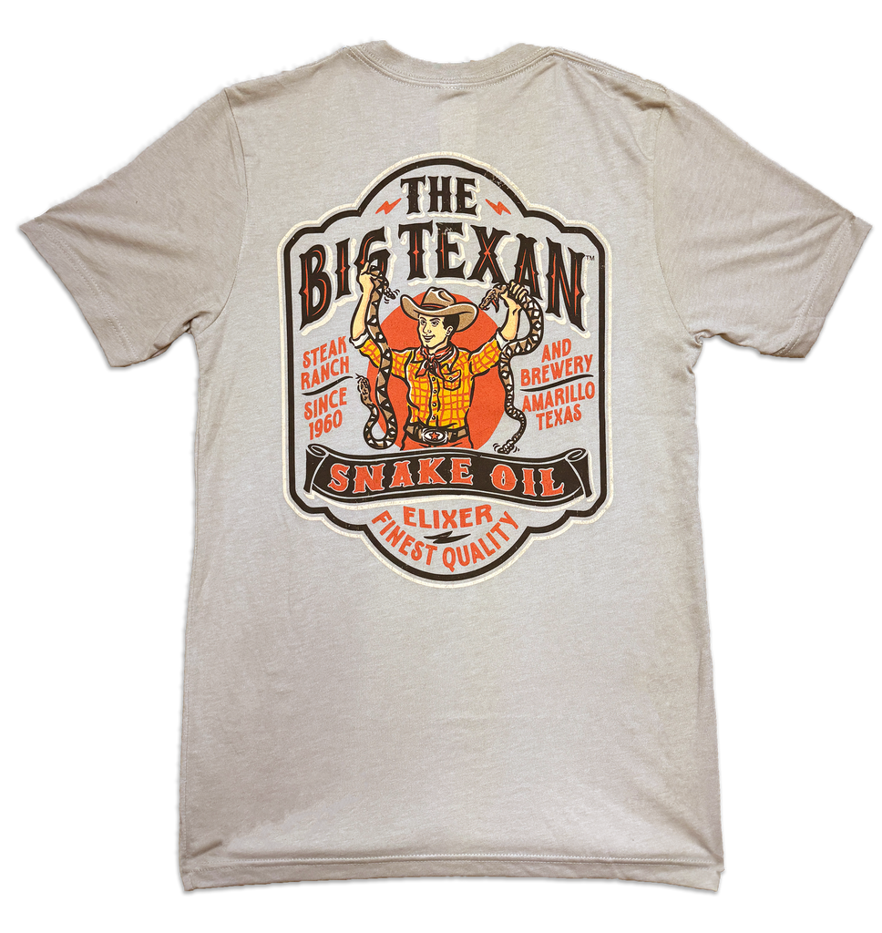 Beige t-shirt back with a graphic reading 'The Big Texan' snake oil featuring a cowboy holding two rattlesnakes in each hand with a banner across his waist that reads' Snake oil'