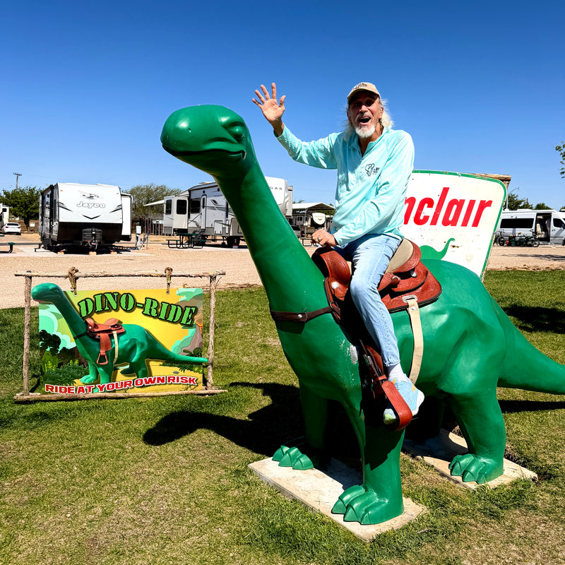 Man riding a green dinosaur statue with RVs and signs in the background