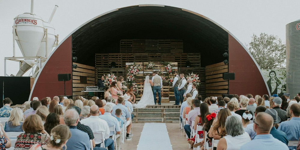 Wedding ceremony taking place in a rustic outdoor setting with a stage and audience.