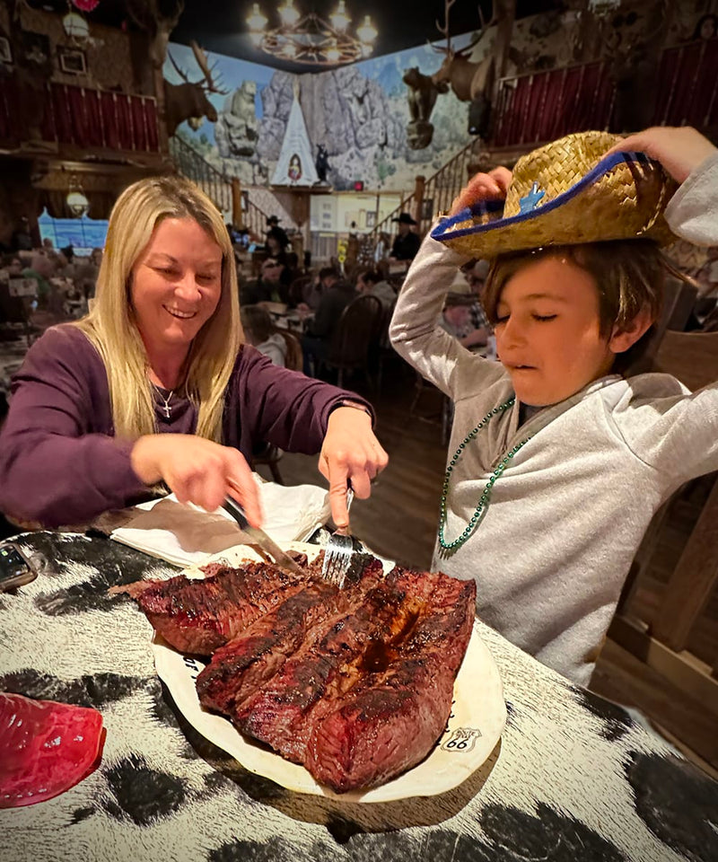 A mom and son at a table with a large plate of meat, one person cutting into it, in a rustic setting.
