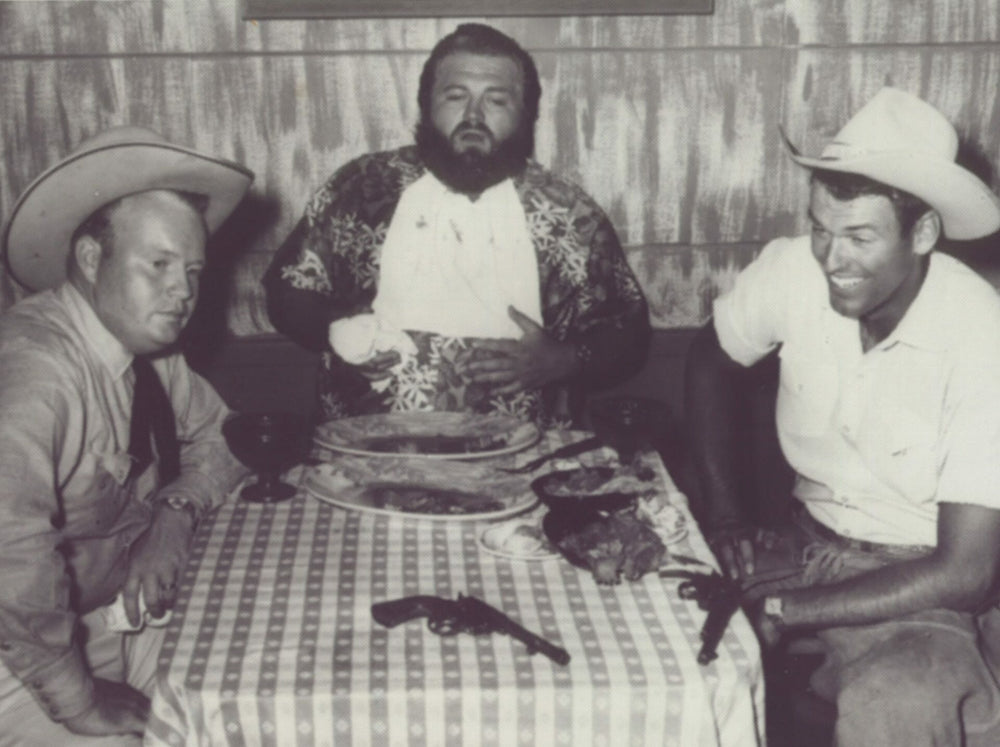 Black and white photo of three men around a table with a gun and hats.